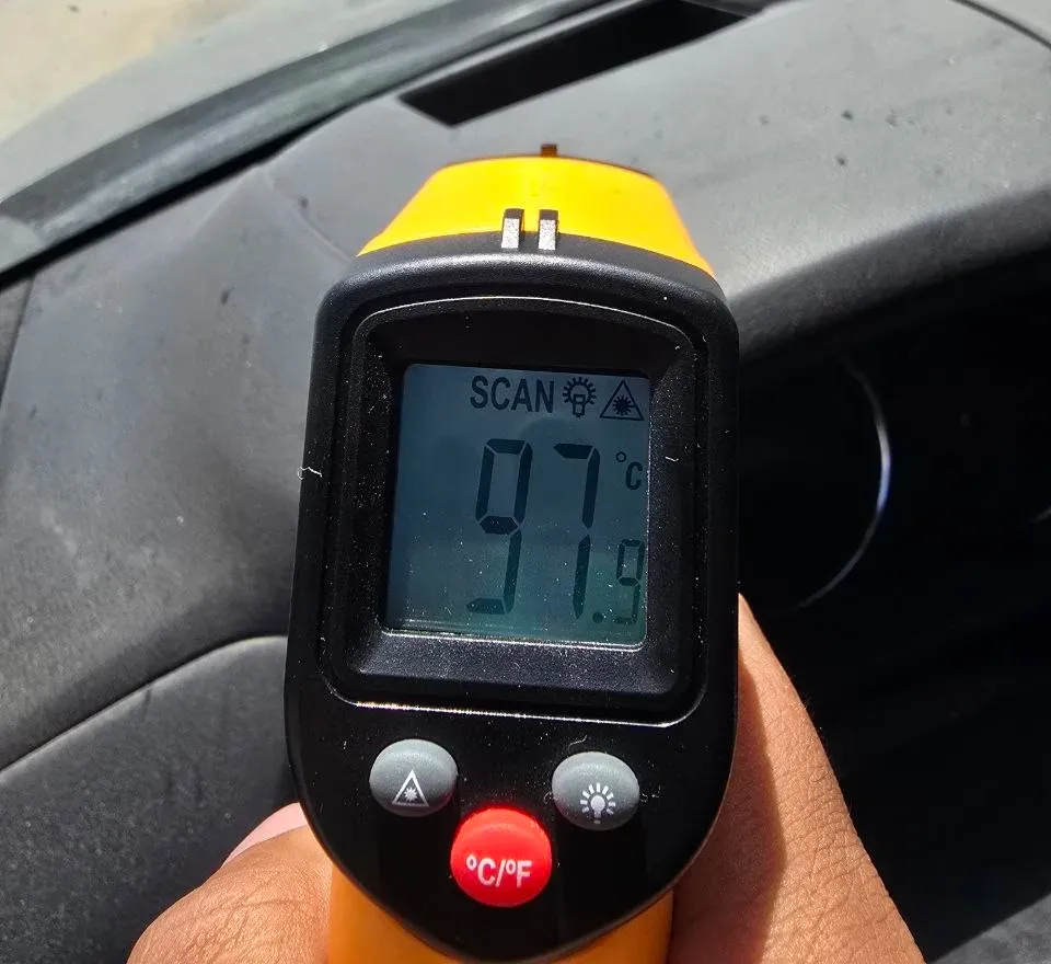 An infrared thermometer reading 97.9C pointed at a car dashboard in direct sun