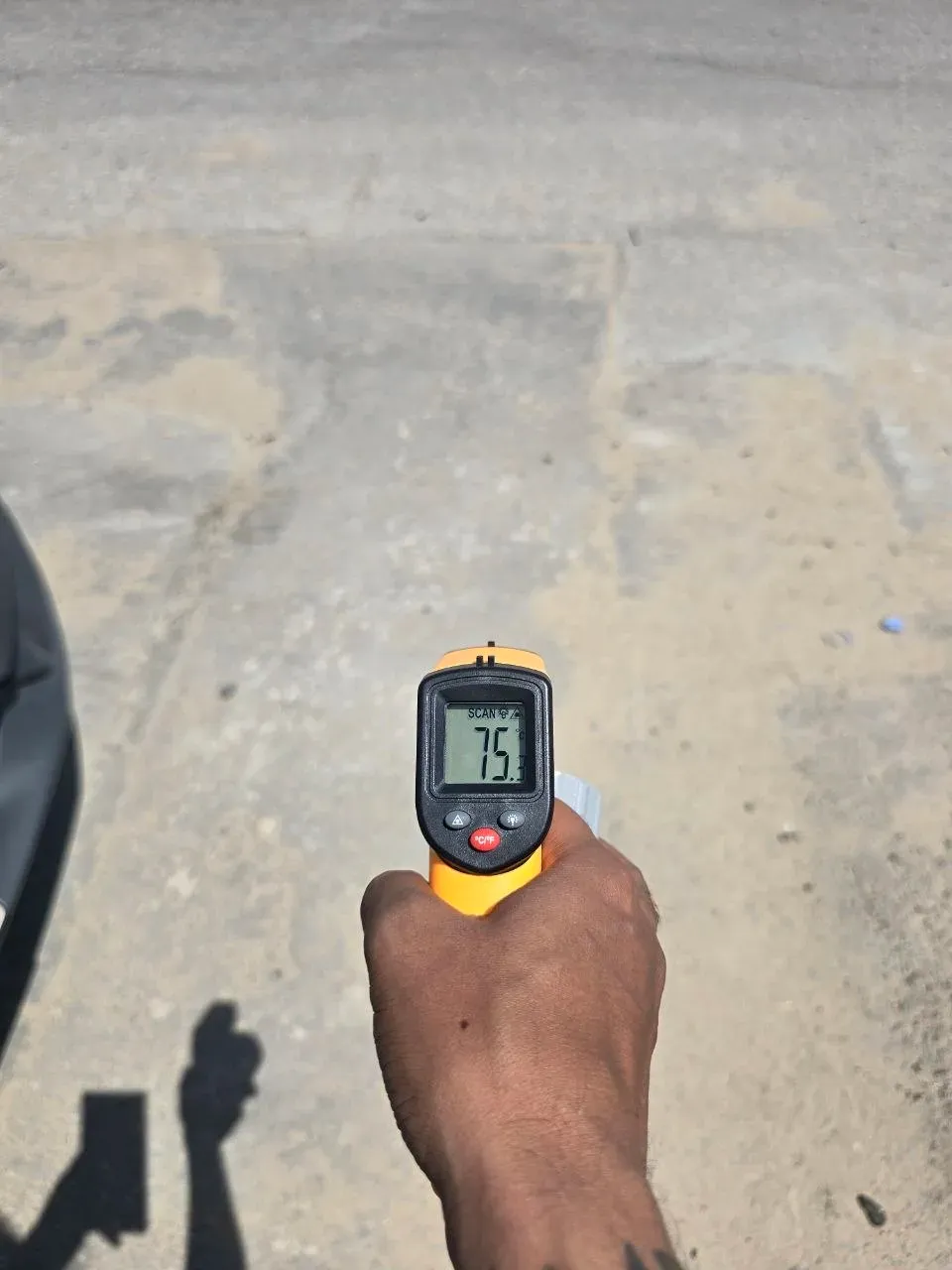 An infrared thermometer reading 75.3C pointed at the asphalt road outside the car